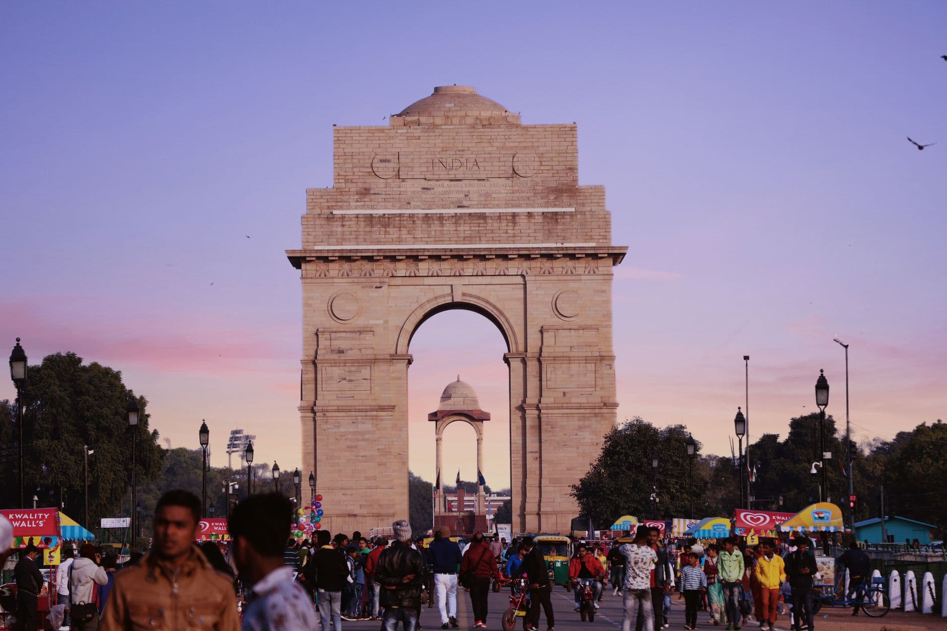 India Gate at dusk under a pink sky, evening crowds and ice-cream carts on the avenue, the empty canopy visible through the arch