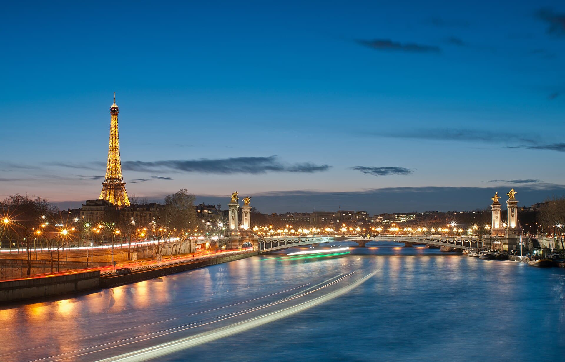 The illuminated Eiffel Tower at night, seen across the Seine beyond the gilded statues of Pont Alexandre III