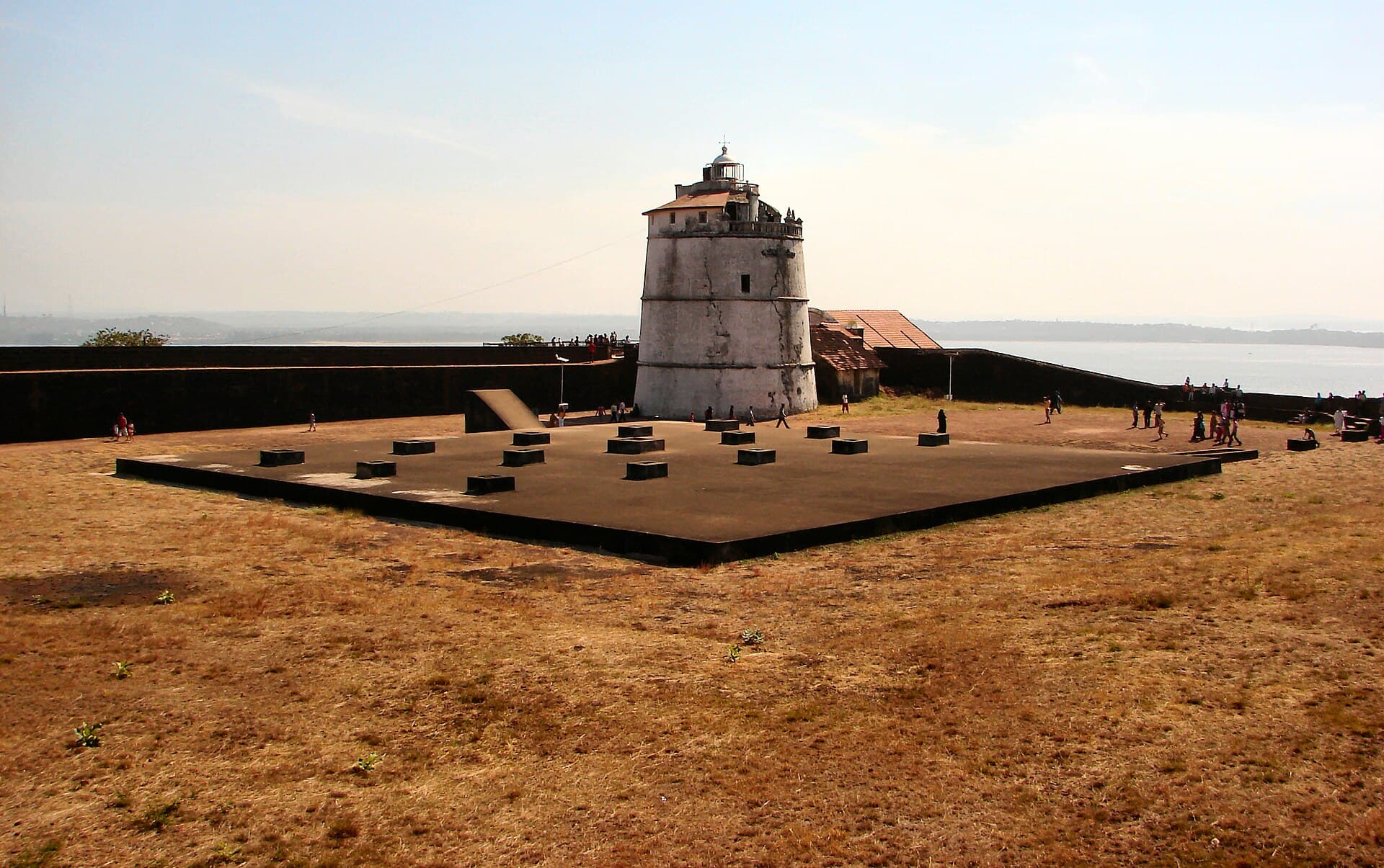 The white four-storey lighthouse of Fort Aguada standing over the fort's laterite ramparts, with the river mouth and sea behind