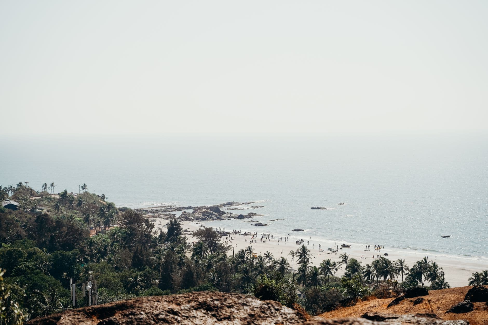 Palm-fringed North Goa coastline seen from a hilltop, with beachgoers on the sand, rocky outcrops in the surf and the Arabian Sea fading to a hazy horizon