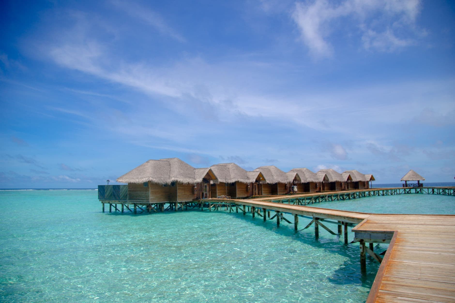 Thatched-roof overwater bungalows on stilts over a clear turquoise Maldivian lagoon under a blue sky
