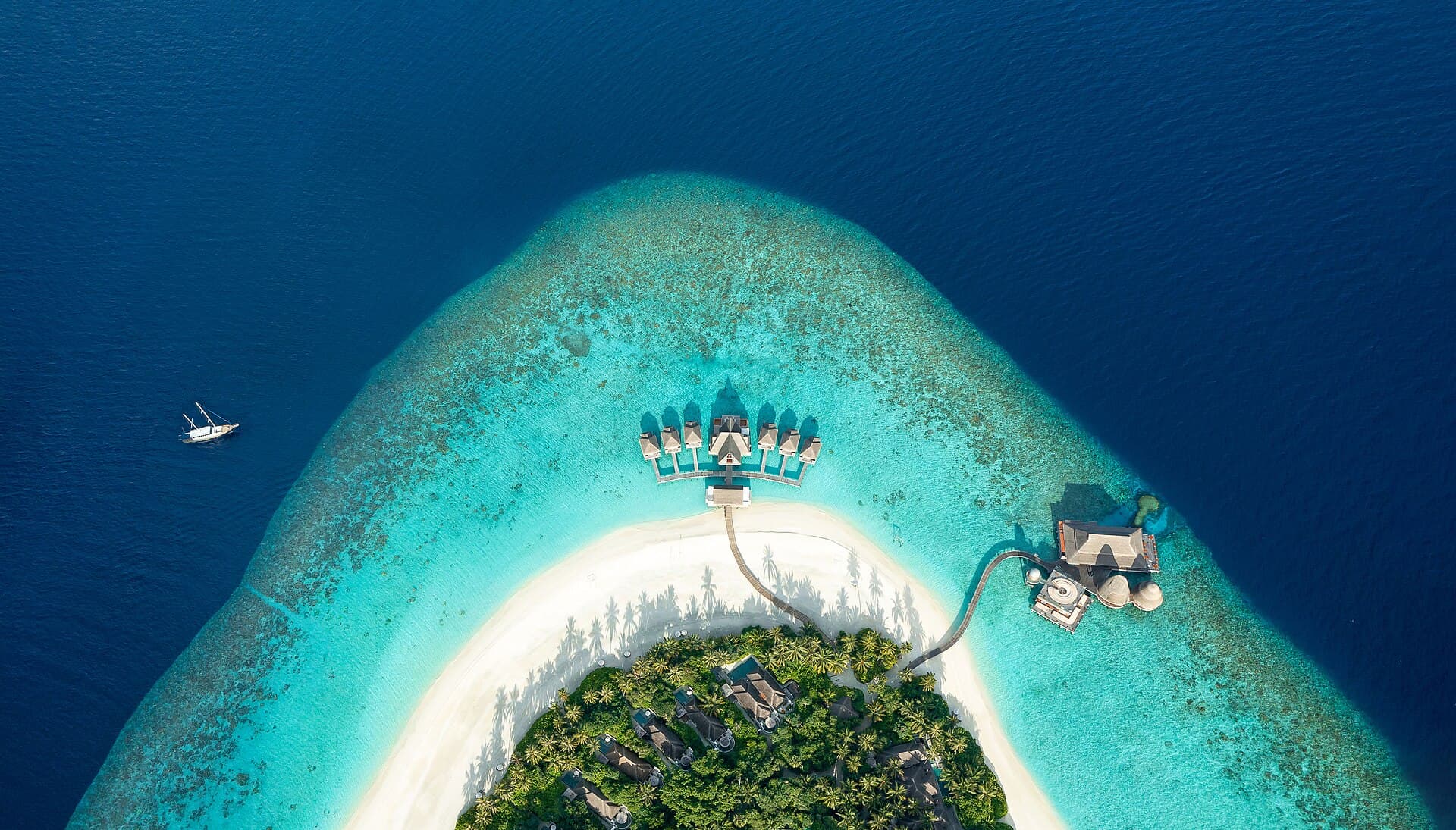 Aerial view of overwater villas fanning out from the tip of a reef island, turquoise shallows giving way to deep blue ocean