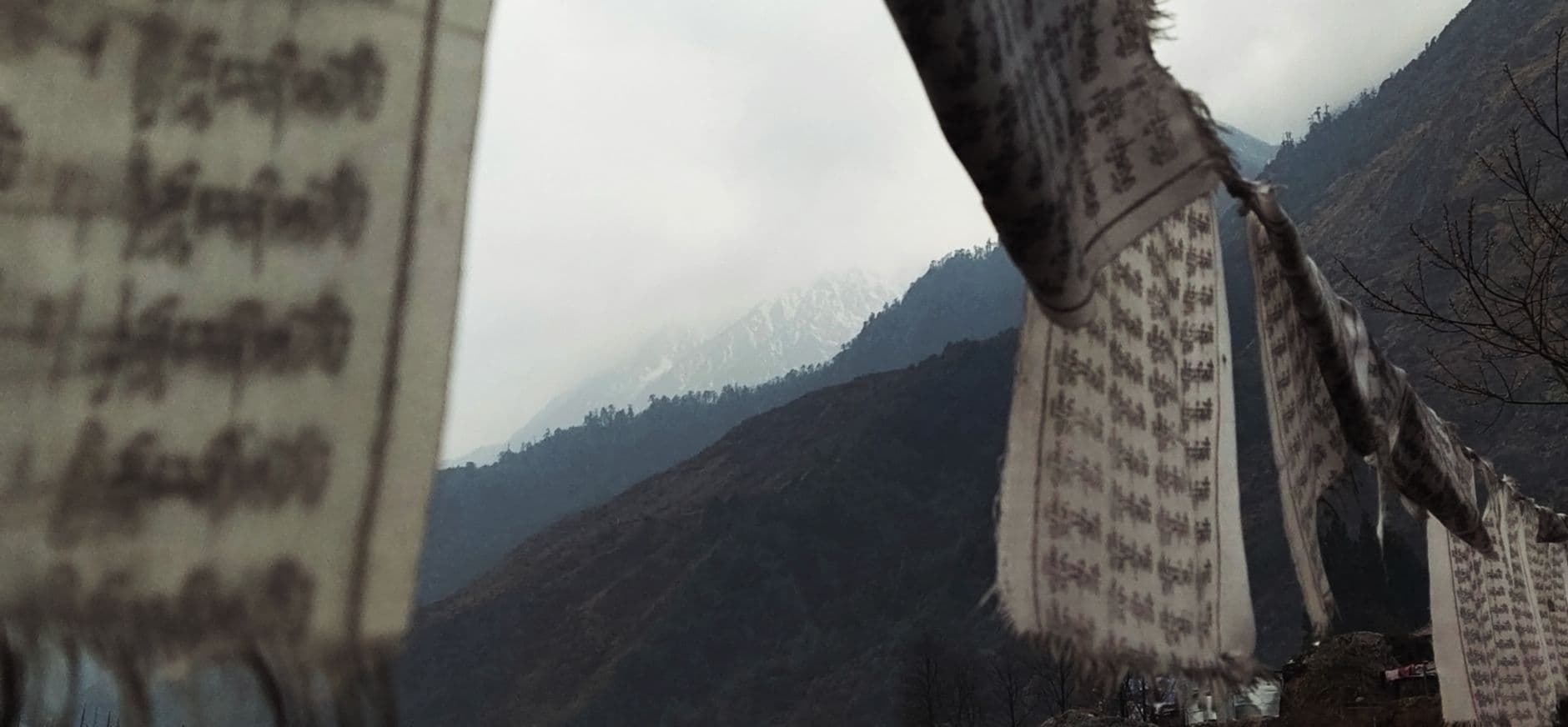 Weathered white prayer flags printed with mantras strung across the frame, misty forested Himalayan ridges and a snow patch behind